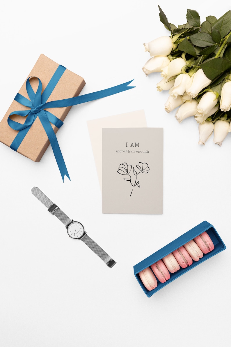 K&ouml;nnte beinhalten: Eine flache Anordnung mit einer Geschenkbox mit blauem Band, einer Karte mit dem Text "I AM more than enough", einer Uhr mit silbernem Armband, einem Strau&szlig; wei&szlig;er Rosen und einer blauen Box mit rosa Macarons.