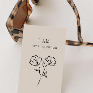 Pode incluir: Um cartão retangular bege com o texto preto I AM more than enough acima de um desenho de linha preta simples de duas flores. O cartão está posicionado parcialmente sob um par de óculos de sol com padrão de tartaruga castanho e preto.