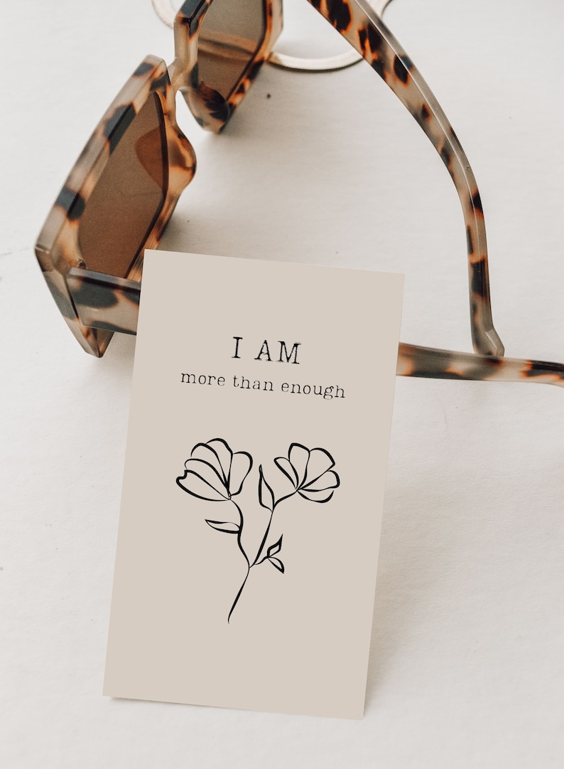 K&ouml;nnte beinhalten: Eine beige Karte mit dem Text "I AM more than enough" und einer einfachen Linienzeichnung von zwei Blumen.