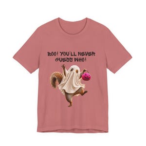 May include: A dusty rose short-sleeve t-shirt featuring a graphic of a squirrel dressed as a ghost, holding a small pink pumpkin bucket. Above the squirrel, the text reads: BOO! YOU'LL NEVER GUESS WHO! The design is centered on the front of the shirt.