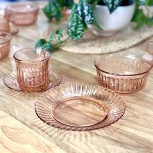 May include: A set of pink glass bowls and cups with a ribbed design. The bowls and cups are arranged on a wooden table with a plant in the background.