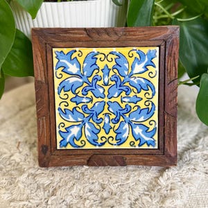May include: Square wooden coaster with a yellow and blue floral tile insert. The tile features a repeating pattern of blue leaves and swirls against a yellow background. The coaster's wooden frame has a dark brown finish and a carved design.