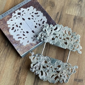 May include: Silver-plated serving pieces with ornate, openwork floral designs. The pieces are resting on a book with the words "ROYAL LIMITED" and a decorative paper doily. The items are on a wooden surface.