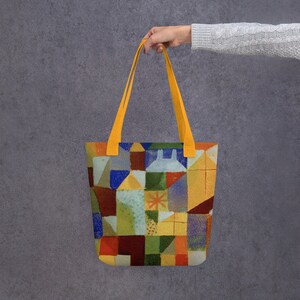 以下が含まれることがあります： カラフルな幾何学模様の黄色いトートバッグ。模様は赤、オレンジ、黄色、緑、青、紫の色合いの正方形、長方形、三角形で構成されています。バッグには黄色のハンドルが2つ付いています。