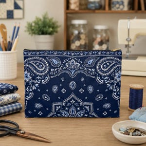 May include: A rectangular zippered pouch featuring a dark navy blue fabric with a white and light blue paisley bandana pattern. The pouch has a gold-toned metal zipper closure along the top edge and a structured, flat-bottomed design.