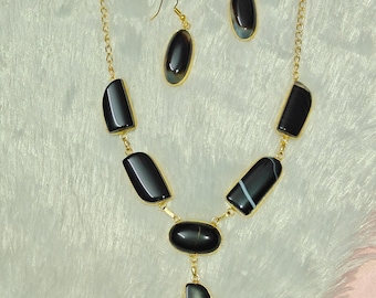 Puede incluir: Un conjunto de collar y pendientes de color dorado con colgantes de piedra negra pulida. El collar tiene una cadena con múltiples piedras rectangulares y ovaladas. Los pendientes tienen piedras ovaladas. El conjunto de joyas se exhibe sobre una superficie blanca texturizada.