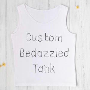 May include: White tank top with the words "Custom Bedazzled Tank" spelled out in small, dark gray rhinestones. The tank top is sleeveless and has a scoop neck. The background is a light wood surface.