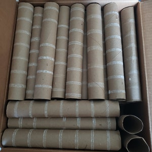 May include: A cardboard box filled with numerous empty brown paper towel rolls. Each roll has white stripes. The rolls are arranged in rows, with some standing upright and others lying flat.