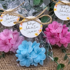 May include: Three small favor boxes with clear cellophane wrappers, each containing a cluster of artificial flowers. The flowers are pink, blue, and white. Each box has a round tag attached with the text "Thank you! Amber & Evan July 20th 2024".
