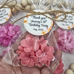May include: Pink flower-shaped favors in clear cellophane bags, tied with twine and a circular tag. The tag reads "Thank you! Journey's 18th Birthday Party Aug. 2025". The favors are surrounded by other similar party favors.