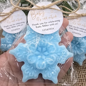 May include: Blue snowflake-shaped soap favor with a white tag that reads "Baby it's cold outside. Thank you for celebrating Baby Miller with us! 12/14/2024".