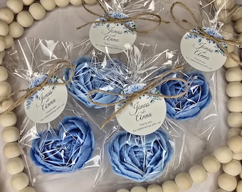 May include: Five blue rose-shaped soaps in clear cellophane bags, tied with twine and tags. The tags read "Jonas & Anna, Thank you for celebrating with us!" The soaps are surrounded by a wooden bead garland.