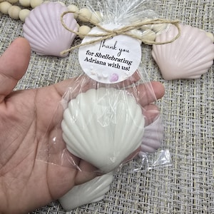 May include: White seashell-shaped soap favors in a clear cellophane bag, tied with twine and a tag that reads "Thank you for Shellebrating Adriana with us!". Additional pink seashell soaps and wooden beads are visible.