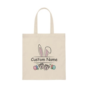 Pode incluir: Uma bolsa de lona bege com um coelho rosa espiando por cima. A bolsa tem o texto "Custom Name" impresso, juntamente com um par de pegadas de coelho e ovos de P&aacute;scoa coloridos.