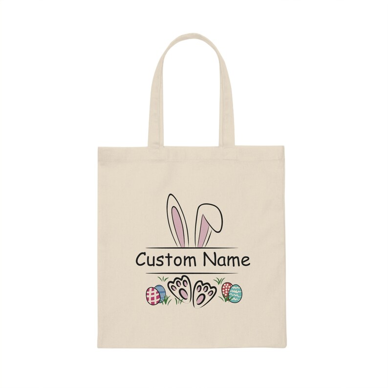 Pode incluir: Uma bolsa de lona bege com um coelho rosa espiando por cima. A bolsa tem o texto "Custom Name" impresso, juntamente com um par de pegadas de coelho e ovos de P&aacute;scoa coloridos.