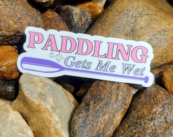May include: A white sticker with pink text that reads "Paddling Gets Me Wet". The sticker features a purple paddle with a white starburst design. The sticker is placed on a pile of brown and tan rocks.