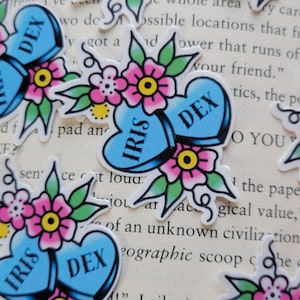 May include: Two blue heart-shaped stickers with the names "Iris" and "Dex" written on them. The stickers are surrounded by green leaves and pink flowers with yellow centers.