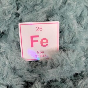 May include: A white and pink periodic table element card for Iron (Fe) with the atomic number 26 and atomic mass 55.845, set against a teal, fluffy background.