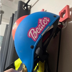 May include: A blue helmet with a pink sticker that says "Beater" on it. The helmet is hanging on a hook.