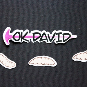 May include: A sticker set featuring the text "OK DAVID" in black and white with a pink arrow. Three worm-like stickers are also included, all on a black background.