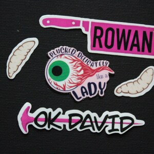 May include: A collection of quirky stickers on a black background. The stickers include a pink cleaver with the name "ROWAN", an eyeball with the text "PLUCKED DELICATELY like a LADY", and the text "OK DAVID" with a pink arrow. Also included are several worm-like stickers.