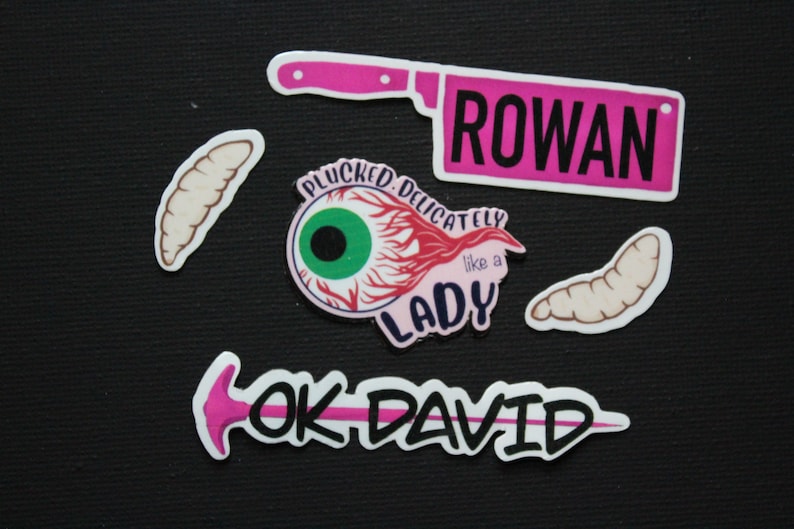 May include: A collection of quirky stickers on a black background. The stickers include a pink cleaver with the name "ROWAN", an eyeball with the text "PLUCKED DELICATELY like a LADY", and the text "OK DAVID" with a pink arrow. Also included are several worm-like stickers.