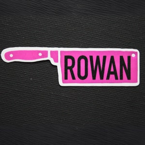 May include: A pink knife-shaped patch with the name "ROWAN" in black block letters. The patch has a white border and is set against a black textured background. The design resembles a kitchen knife.