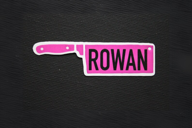 May include: A pink knife-shaped patch with the name "ROWAN" in black block letters. The patch has a white border and is set against a black textured background. The design resembles a kitchen knife.