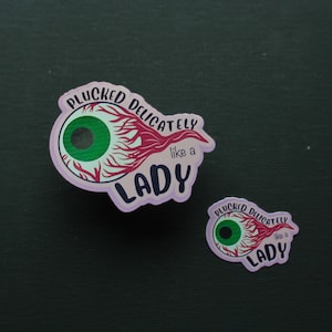May include: Two stickers with an eye design and the text "PLUCKED DELICATELY like a LADY". The eye has a green iris, red veins, and a light purple outline. One sticker is larger than the other.