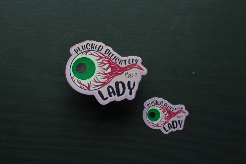May include: Two stickers with an eye design and the text "PLUCKED DELICATELY like a LADY". The eye has a green iris, red veins, and a light purple outline. One sticker is larger than the other.