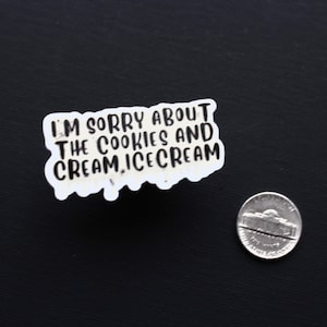 May include: A white sticker with the text "I'M SORRY ABOUT THE COOKIES AND CREAM, ICE CREAM" in black lettering. A nickel is next to the sticker on a black surface.