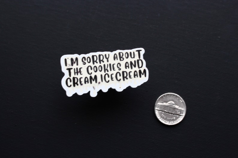 May include: A white sticker with the text "I'M SORRY ABOUT THE COOKIES AND CREAM, ICE CREAM" in black lettering. A nickel is next to the sticker on a black surface.