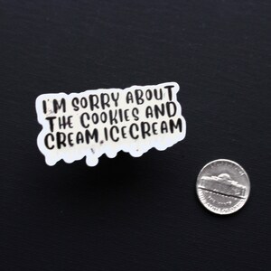 May include: A white sticker with the text "I'M SORRY ABOUT THE COOKIES AND CREAM, ICE CREAM" in black lettering. A nickel is next to the sticker on a black surface. The sticker has a unique, irregular shape.