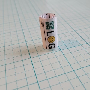 Puede incluir: Un tubo de papel blanco con la palabra "LOG" impresa en letras negras. Una cara sonriente amarilla está impresa sobre la palabra "LOG". El tubo está sobre una superficie blanca cuadriculada.