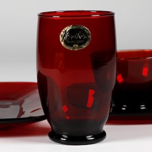 May include: A red glass dinnerware set, including a rectangular plate, a tall tumbler, a teacup, and a saucer. The glass has a dark red hue with a black base. A gold sticker is on the tumbler. The set is designed for serving food and beverages.