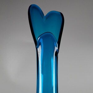 May include: A tall, elegant, turquoise-blue glass vase. The vase has a slender, elongated body that flares out at the base. The top features a heart-shaped design. The glass has a glossy finish, reflecting light.