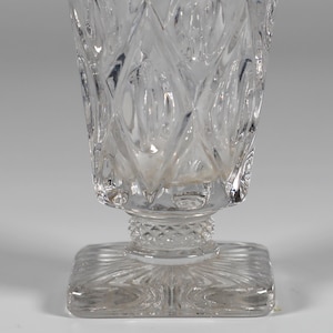 Op de afbeelding: Transparante, geslepen glazen beker met een vierkante basis en een ruitpatroon. Het glas heeft een cilindrische vorm met een decoratieve band aan de bovenkant en een gedetailleerde basis. Het ontwerp is symmetrisch en het glas is transparant.