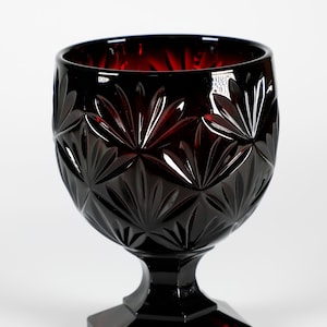 Può includere: Un calice di vetro rosso scuro con un design in vetro tagliato. Il calice ha una coppa larga e un gambo corto con una base esagonale.