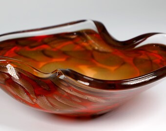 May include: A red and gold glass bowl with a wavy, irregular shape. The bowl has a clear glass rim and a swirling pattern of red and gold.