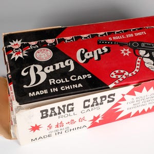 May include: A red and black box of Bang Caps roll caps for repeating pistols. The box has a picture of a toy pistol and the text "Bang Caps Roll Caps Made in China" on the front.