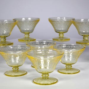 Puede incluir: Un conjunto de nueve copas de postre de vidrio teñido de amarillo. Cada copa tiene un cuenco acampanado con un delicado diseño floral y se apoya en un tallo corto y redondo. Las copas están dispuestas sobre soportes de acrílico transparente.