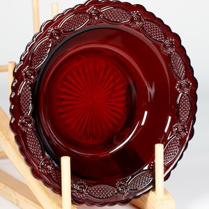 May include: A deep red glass plate featuring a decorative scalloped edge with embossed floral and geometric patterns. The plate's centre has a sunburst design. It is displayed on a wooden plate stand.