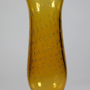 May include: Amber-colored glass vase with a flared top and a rounded base. The vase has a textured surface with small, irregular bubbles and etched vertical lines near the top. The base is a solid, circular shape. A label is visible at the base.