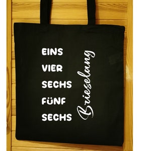 Könnte beinhalten: Schwarze Einkaufstasche mit weißem Text, der "Eins Vier Sechs Fünf Sechs" und "Briessolong" auf der Seite lautet.