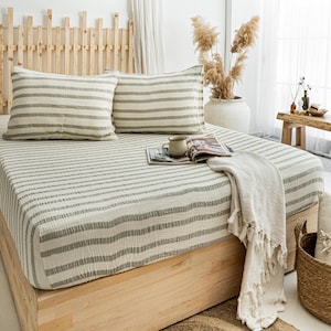 May include: A bed with a light wood frame and headboard, covered with a white and gray striped sheet set. Two matching pillowcases are on the bed. A white fringed throw blanket and a woven basket are also visible.