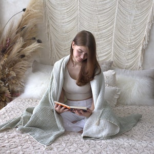 May include: A woman sits on a bed with a white crocheted headboard, reading a book while wrapped in a light green waffle weave blanket. The bed is covered in a white crocheted bedspread and has several white pillows.