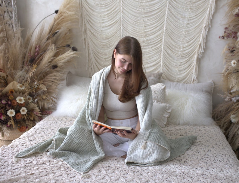May include: A woman sits on a bed with a white crocheted headboard, reading a book while wrapped in a light green waffle weave blanket. The bed is covered in a white crocheted bedspread and has several white pillows.
