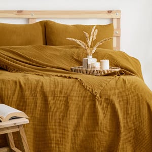 May include: A mustard yellow muslin bedspread and two matching pillowcases with a crinkled texture and fringed edges. A woven tray sits on the bed, holding a small vase with dried grasses and two white candles, next to a wooden stool with a book.