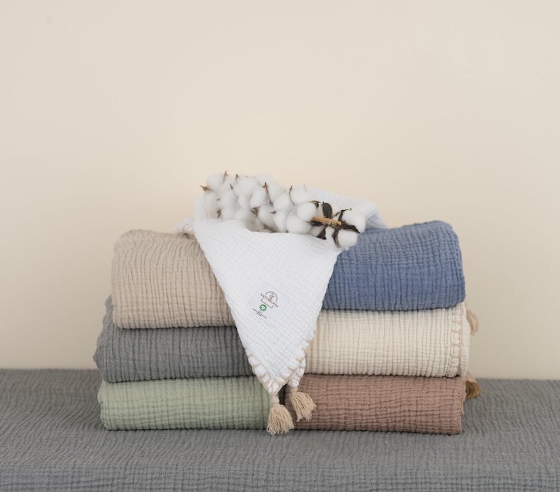 May include: A stack of five soft, textured blankets in various colors, including beige, gray, green, and blue. The blankets are arranged in a pyramid shape with a sprig of cotton on top.