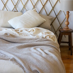 May include: A bed featuring a bamboo headboard, cream-coloured pillows, and a textured blanket in beige and cream hues with a fringed edge. A wooden lamp and stool are positioned beside the bed, creating a cosy bedroom setting.